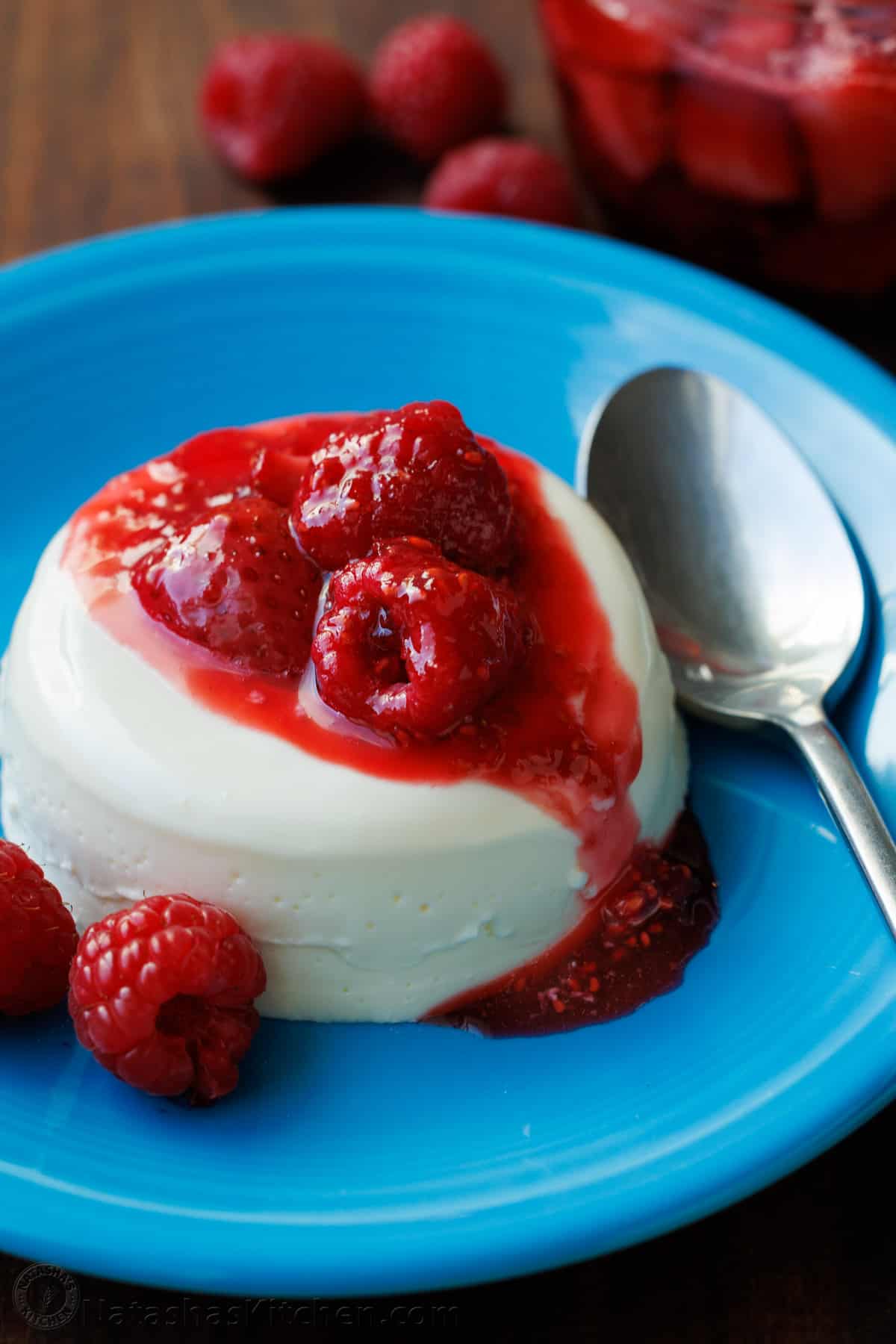 Panna Cotta with berry sauce drizzled on top, sitting on a blue plate with a spoon. 
