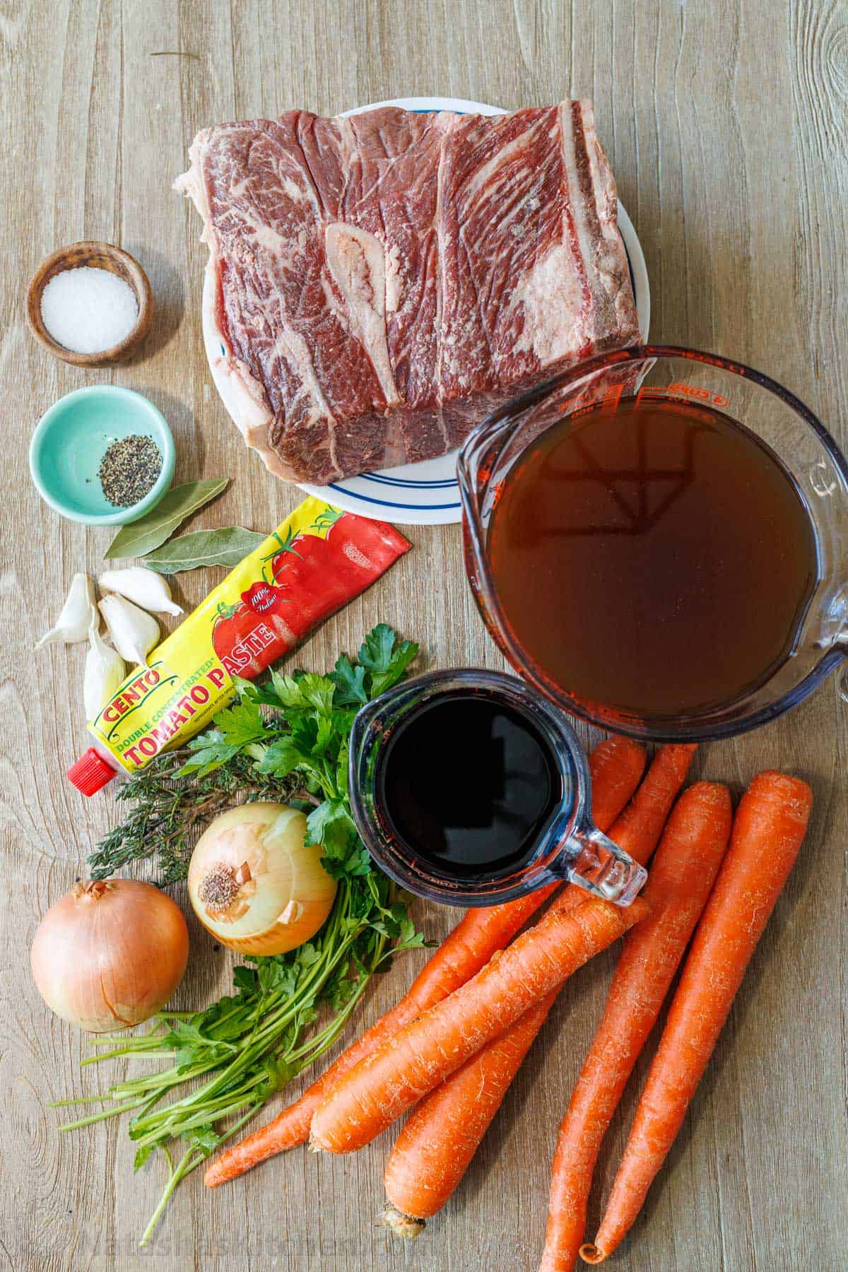 ingredients for cooking beef in the oven including chuck shoulder steak, salt, pepper, garlic, bay leaves, tomato paste, onion, parsley, thyme, carrots, red wine and beef broth