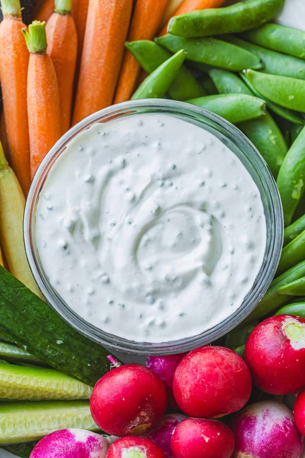 Ranch dip in a clear bowl surrounded by raw vegetables like carrots, cucumbers, snap peas and radishes