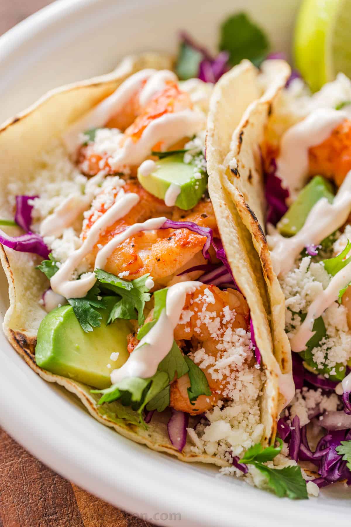 My easy Shrimp Tacos are loaded with shrimp, cabbage slaw, cotija cheese, and the BEST garlic-lime crema sauce. It's the only recipe you need! Side profile of tacos laying on platter loaded with shrimp and drizzled with garlic-lime crema sauce