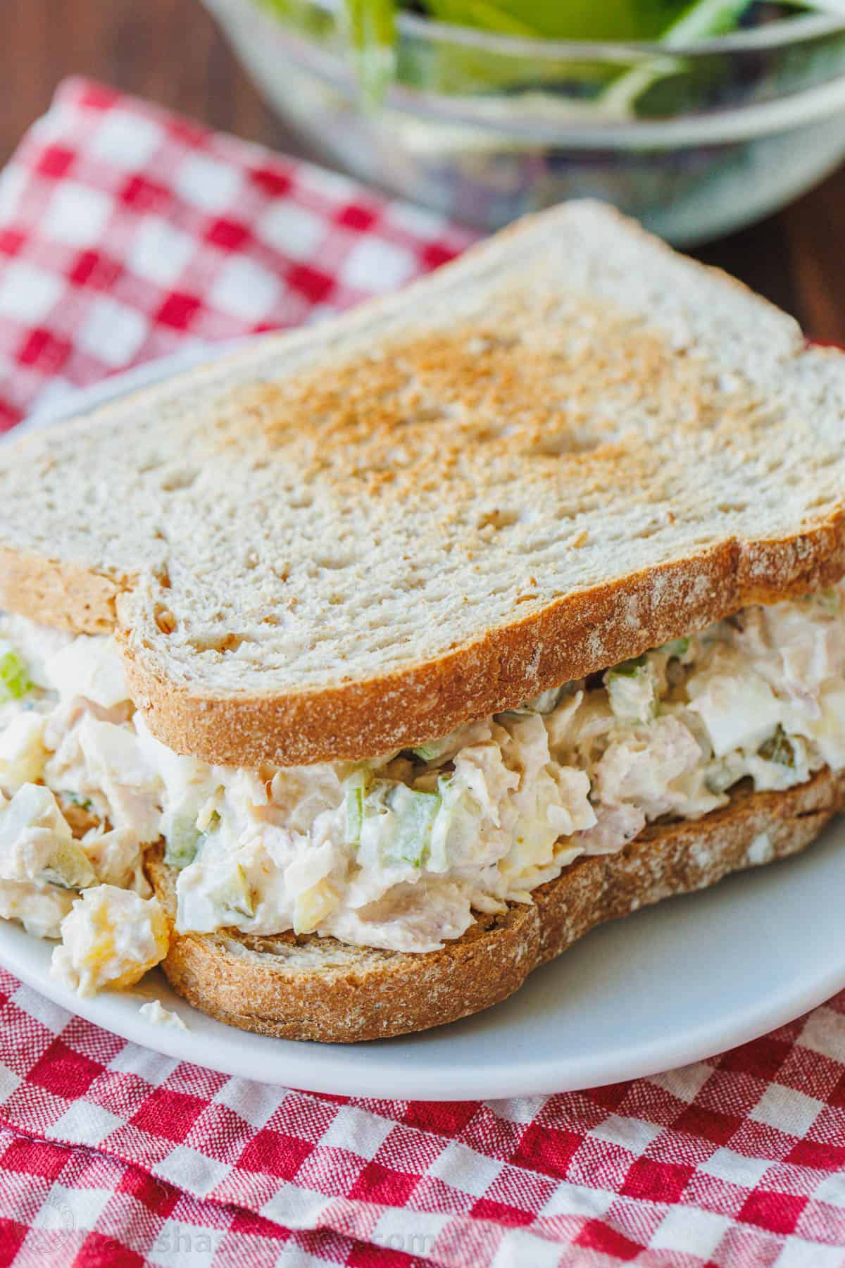 A classic tuna salad recipe served on a sandwich on a white plate, with a red and white checked cloth underneath.